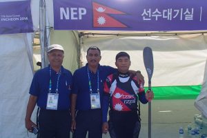 Nim Bahadur Magar stands with Nepalese delegates Kumar Ranabhat and Surya Joshi in official Nepal team uniforms beside event signage displaying the Nepal flag at the 2014 Asian Games in Incheon, South Korea.