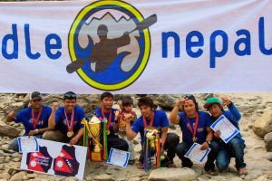 Young Kailash Magar stands with his father, Nim Bahadur Magar, and the Paddle Nepal team as they celebrate a kayaking competition victory with trophies.